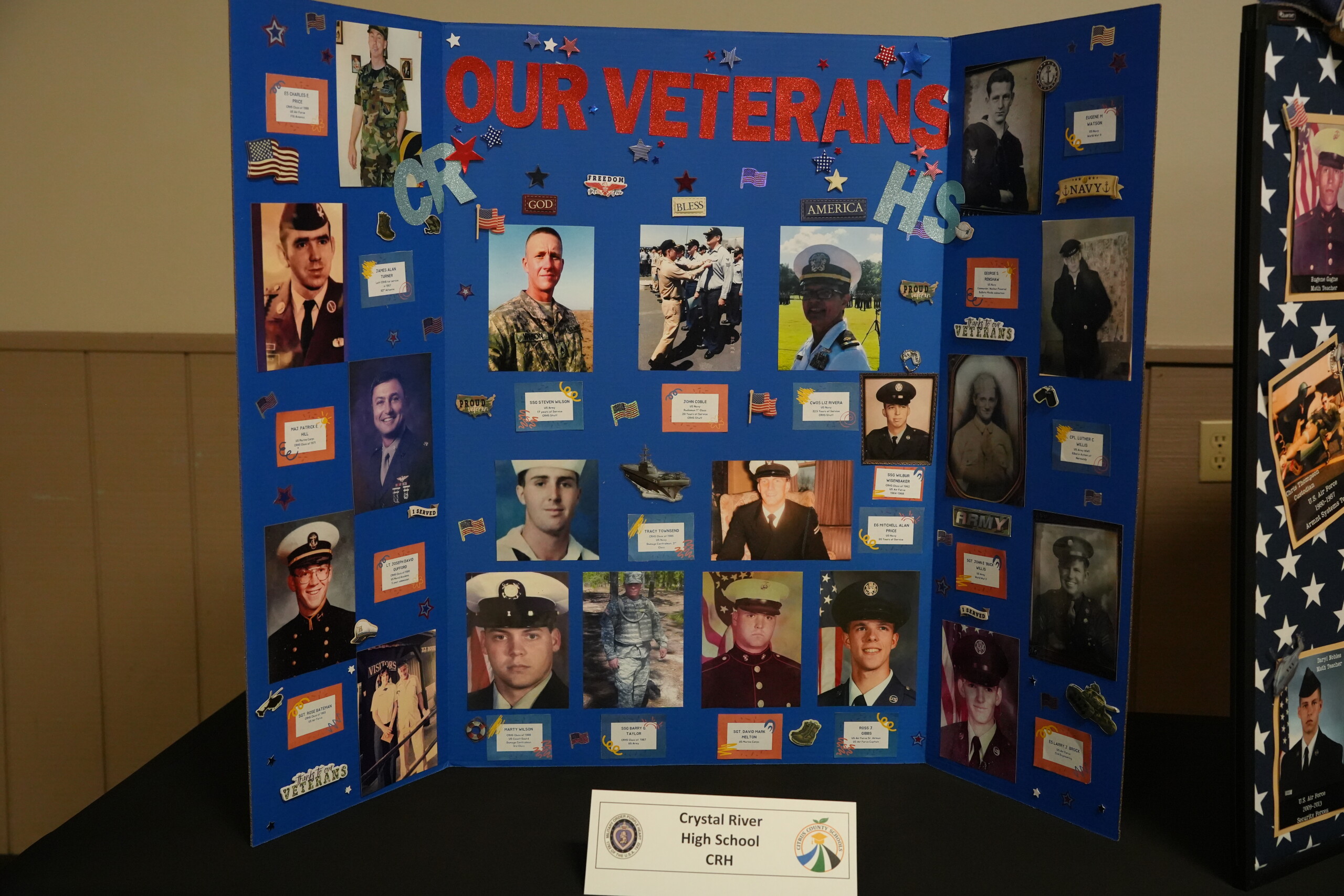 A blue tri-fold display board titled 'OUR VETERANS CR HS' is covered with photographs of service members in uniform.